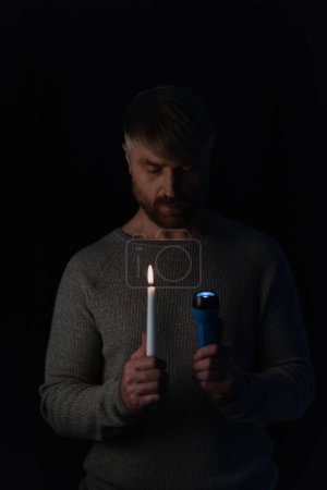 Mann in der Dunkelheit durch Stromabschaltung verursacht hält glühende Taschenlampe und entzündete Kerze isoliert auf schwarz