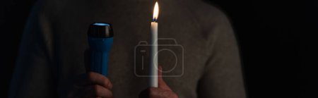 cropped view of man holding electric flashlight and lit candle isolated on black, banner
