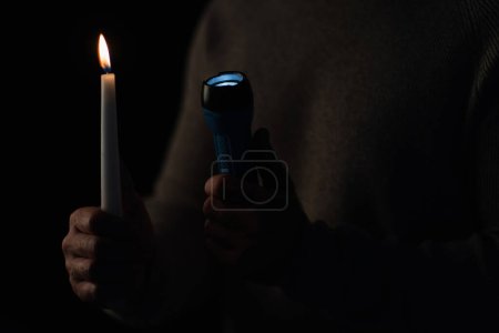 burning candle and glowing flashlight in hands of cropped man isolated on black