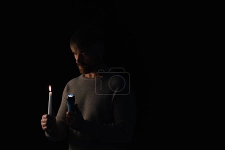 man in darkness holding glowing flashlight and burning candle isolated on black