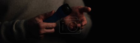 Téléchargez les photos : Vue partielle de l'homme tenant une lampe de poche électrique pendant la panne d'électricité isolé sur noir, bannière - en image libre de droit