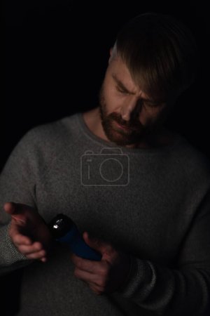 Téléchargez les photos : Adulte homme regardant lampe de poche électrique pendant panne de courant isolé sur noir - en image libre de droit
