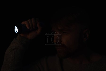 man looking away in darkness while holding bright flashlight isolated on black