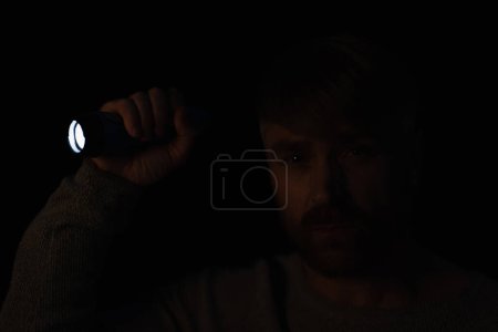 hombre en la oscuridad mirando a la cámara mientras sostiene la linterna brillante aislado en negro