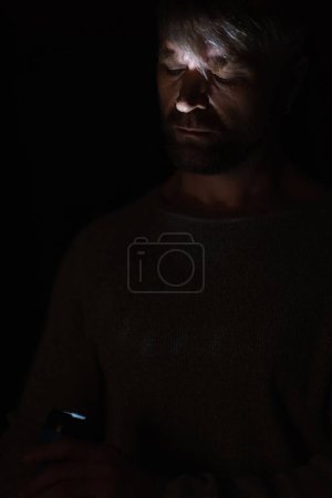 Téléchargez les photos : Homme avec lampe de poche éclatante allumant son visage pendant la panne d'électricité isolé sur noir - en image libre de droit