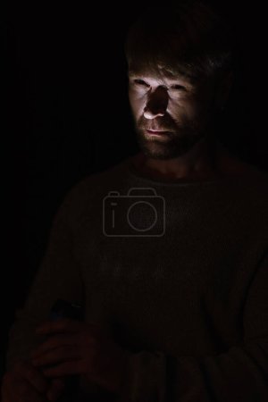 man lighting on face with flashlight while standing in darkness isolated on black