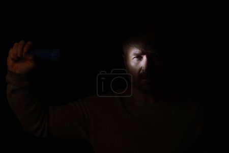 man in darkness lighting on face with flashlight and looking at camera isolated on black