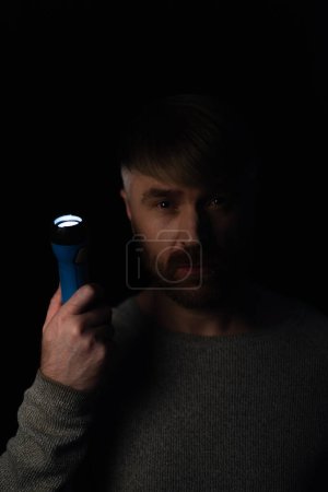 Téléchargez les photos : Homme avec lampe de poche lumineux regardant la caméra pendant l'arrêt de l'électricité isolé sur noir - en image libre de droit