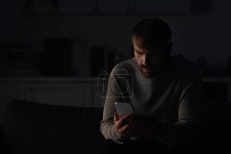 Mann sitzt bei Stromausfall in dunkler Küche und nutzt Handy