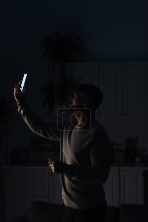 Foto de Hombre de pie en la cocina oscura con teléfono inteligente en la mano levantada y en busca de conexión durante el apagón de energía - Imagen libre de derechos