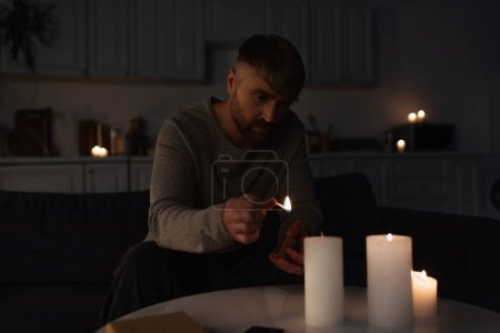 Mann hält brennendes Streichholz in der Hand, während er Kerzen in dunkler Küche bei Stromausfall anzündet