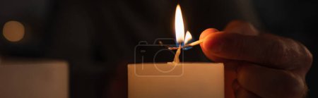 partial view of man lighting candle with burning match during electricity blackout on black background, banner