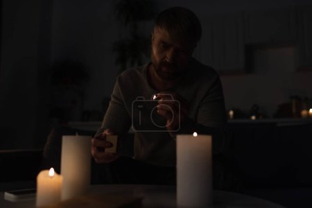 Mann hält brennendes Streichholz neben brennenden Kerzen, während er während eines Stromausfalls in der Küche sitzt