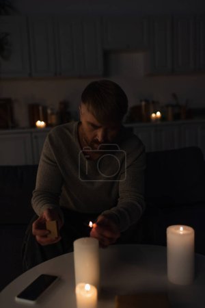 Téléchargez les photos : Homme assis dans la cuisine sombre pendant panne d'énergie et d'allumer des bougies - en image libre de droit