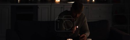 Foto de Hombre con fósforo encendido sentado en la cocina cerca de velas durante el apagón de energía, pancarta - Imagen libre de derechos