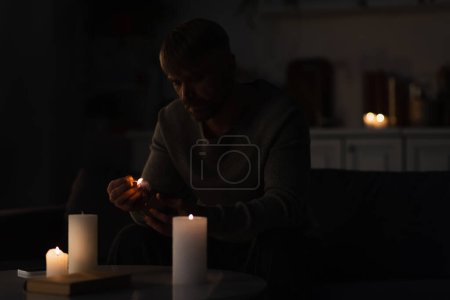 Téléchargez les photos : Homme avec allumette allumette assis près des bougies et réserver sur la table pendant la panne d'énergie - en image libre de droit