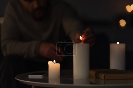 partial view of man in darkness lighting candle near book and smartphone on table 