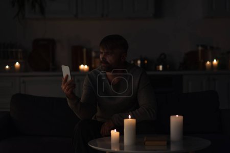 man looking at mobile phone while sitting in dark kitchen near burning candles 