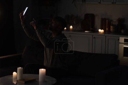 Mann sitzt in Dunkelheit neben brennenden Kerzen und fängt Handy-Verbindung mit Smartphone ein