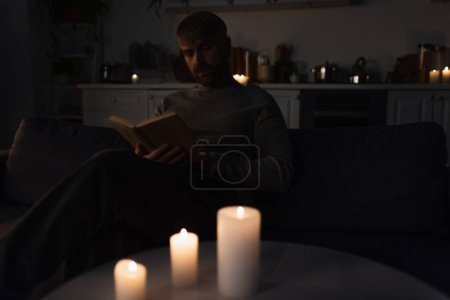 man sitting in kitchen during energy blackout and reading book near lit candles 