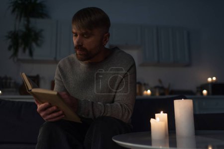 Mann liest Buch, während er bei Stromausfall in dunkler Küche neben brennenden Kerzen sitzt