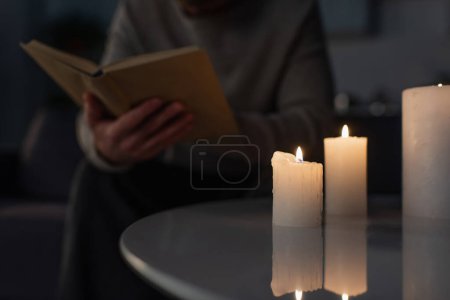 partial view of blurred man reading book in darkness near burning candles on table