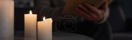 selektiver Fokus brennender Kerzen in der Nähe des zugeschnittenen Mannes, der in der Dunkelheit Buch liest, auf verschwommenem Hintergrund, Banner