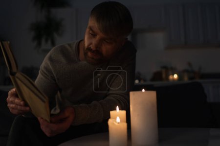 Mann liest Buch, während er in dunkler Küche neben brennenden Kerzen sitzt 