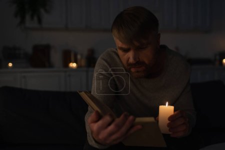 Mann mit brennender Kerze beim Lesen in dunkler Küche bei Stromausfall