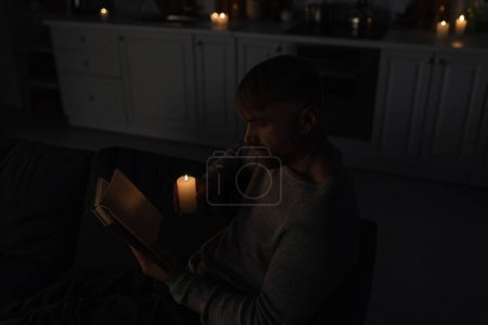 Téléchargez les photos : Vue grand angle du livre de lecture de l'homme pendant l'arrêt de l'électricité dans la cuisine avec des bougies allumées - en image libre de droit
