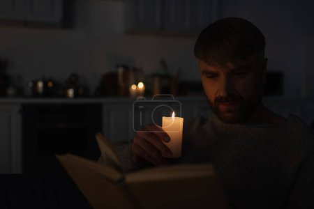 Téléchargez les photos : Homme tenant bougie allumée tout en lisant le livre dans la cuisine sombre pendant l'arrêt de l'électricité - en image libre de droit