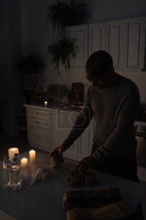 mężczyzna gospodarstwa konserwy żywności w pobliżu butelkowanej wody i świece w kuchni podczas zaciemnienia zasilania