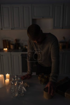 Téléchargez les photos : Homme debout dans la cuisine sombre près du stock de nourriture en conserve avec de l'eau en bouteille et des bougies - en image libre de droit