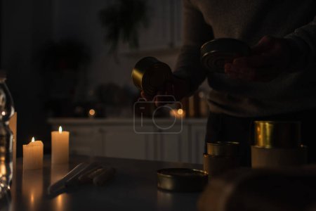 Téléchargez les photos : Vue partielle de l'homme tenant de la nourriture en conserve près des bougies dans la cuisine pendant l'arrêt de l'électricité - en image libre de droit