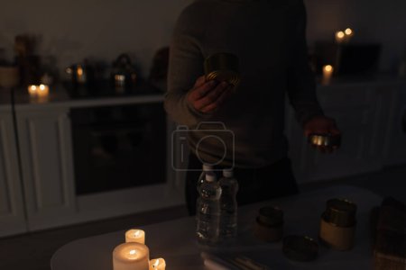 Téléchargez les photos : Vue recadrée de l'homme avec de la nourriture en conserve près de l'eau embouteillée et des bougies allumées dans la cuisine sombre - en image libre de droit
