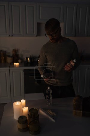 hombre con anteojos sosteniendo agua embotellada cerca de reserva de comida enlatada y velas sobre mesa en cocina oscura