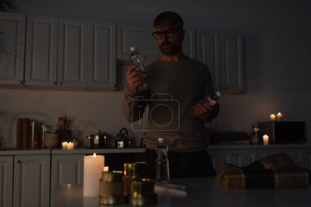 mężczyzna w okularach trzyma butelkowaną wodę przy stole z konserwami i ciepłym kocem w ciemnej kuchni