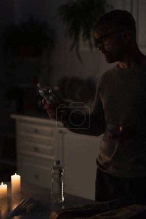 hombre sosteniendo agua embotellada cerca de velas encendidas en la cocina oscura durante el apagado de la electricidad