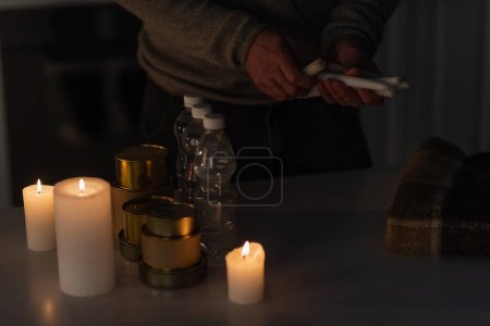 Téléchargez les photos : Vue partielle de l'homme avec des bougies près de la table avec couverture chaude et réserve d'eau et de nourriture en conserve - en image libre de droit