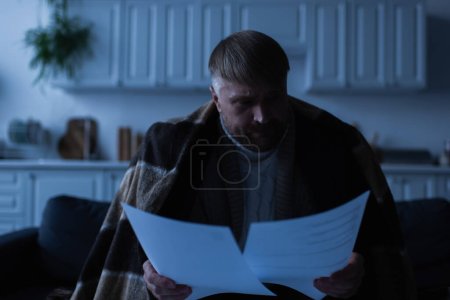 Unzufriedener Mann schaut sich Rechnungen an, während er während des Stromausfalls in der Küche unter einer Decke sitzt