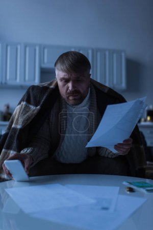 Téléchargez les photos : Homme inquiet sous couverture chaude tenant smartphone et factures au crépuscule à la maison - en image libre de droit