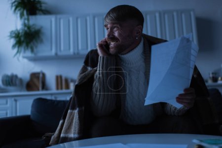 hombre nervioso y congelado hablando en el teléfono inteligente mientras sostiene las facturas de pago en el crepúsculo 