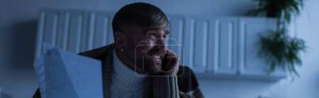 Photo for Nervous man in warm blanket holding invoices while talking on smartphone during energy shutdown, banner - Royalty Free Image