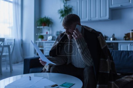 irritated man looking at payment bills and talking on smartphone while sitting under blanket during energy blackout