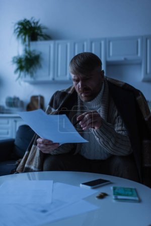 Verspannter Mann zeigt in Küche auf Geldscheine und Smartphone