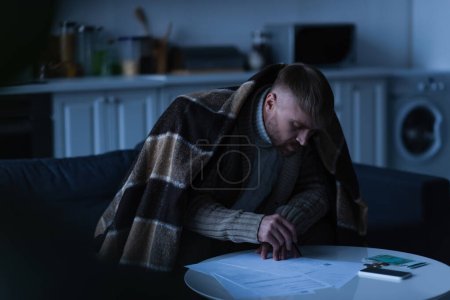 Mann sitzt unter Decke und schaut bei Stromausfall auf Zahlungsrechnungen