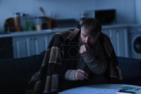 eingefrorener Mann schaut weg, während er unter einer Decke in der Nähe von Rechnungen und Geld während eines Stromausfalls sitzt