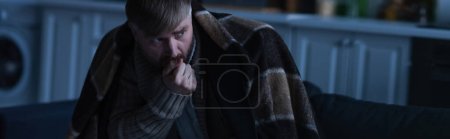 hombre congelado sosteniendo la mano cerca de la cara mientras está sentado bajo una manta caliente y mirando hacia otro lado en el crepúsculo, pancarta