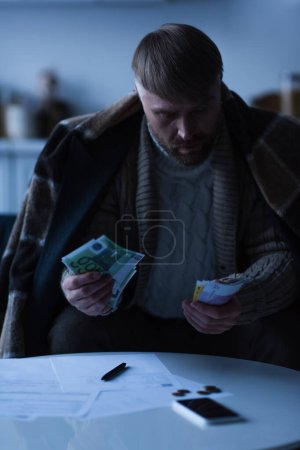 man under warm blanket counting euro banknotes near payment bills during energy shutdown