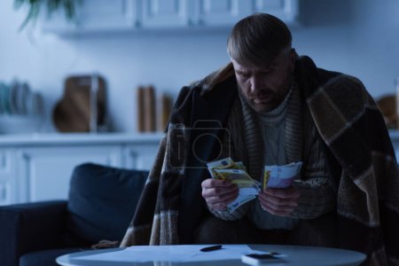 Foto de Hombre sentado debajo de la manta y contando dinero cerca de facturas durante el apagón de energía - Imagen libre de derechos